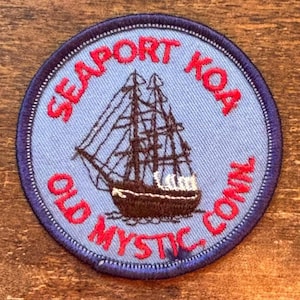Pode incluir: Um patch circular com fundo azul claro e borda azul escuro. O patch apresenta uma ilustração de veleiro em preto e branco. As palavras "SEAPORT KOA" e "OLD MYSTIC CONN" estão em vermelho, arqueadas na parte superior e inferior do patch.