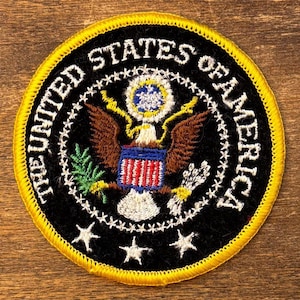 May include: Circular patch featuring the United States of America's seal. The patch has a black background with white text and a yellow border. The seal includes an eagle, stars, and the text "THE UNITED STATES OF AMERICA."