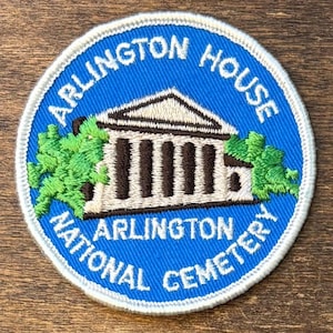 May include: A circular embroidered patch with a bright blue background. It features a white building with dark brown columns, flanked by green trees. The text reads ARLINGTON HOUSE at the top and ARLINGTON NATIONAL CEMETERY at the bottom.
