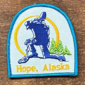 May include: Embroidered patch with a blue figure kneeling, a yellow arc, and green trees on a light blue background. The text "Hope, Alaska" is in yellow. The patch has a blue border.