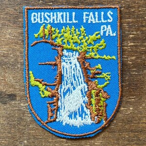 Peut inclure: Écusson brodé avec un fond bleu portant l'inscription "Bushkill Falls PA". L'écusson représente une cascade avec des arbres et une bordure marron. Le motif comprend des couleurs blanches, vertes et marron.