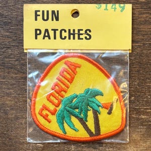 May include: A triangular embroidered patch in a clear plastic bag with a yellow header card labeled FUN PATCHES. The patch features a yellow background, an orange border, the word FLORIDA in orange, and two green palm trees with brown trunks.