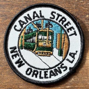 May include: Circular embroidered patch with a green and gold streetcar design, and the text "CANAL STREET NEW ORLEANS LA". The patch has a white background and a black border, representing a New Orleans landmark.