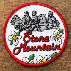 May include: Embroidered patch with a red border and a white interior. The patch features a depiction of figures on horseback, the text "Stone Mountain" in red script, and white flowers with yellow centers.