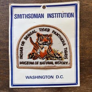 May include: A vintage patch from the Smithsonian Institution featuring an embroidered tiger. The patch is on a white background with blue borders and text that reads "Indian or Bengal Tiger Panthera Tigris" and "Museum of Natural History".