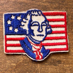 May include: Embroidered patch depicting a historical figure against the backdrop of the American flag. The patch features a blue and white portrait, with the flag's red and white stripes and blue star field.