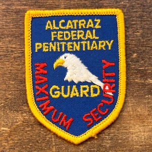 以下が含まれることがあります： 「ALCATRAZ FEDERAL PENITENTIARY」と「MAXIMUM SECURITY GUARD」の文字が入った青、黄、赤のパッチ。黄色い嘴を持つ白いワシが描かれています。パッチには黄色の縁取りがあります。