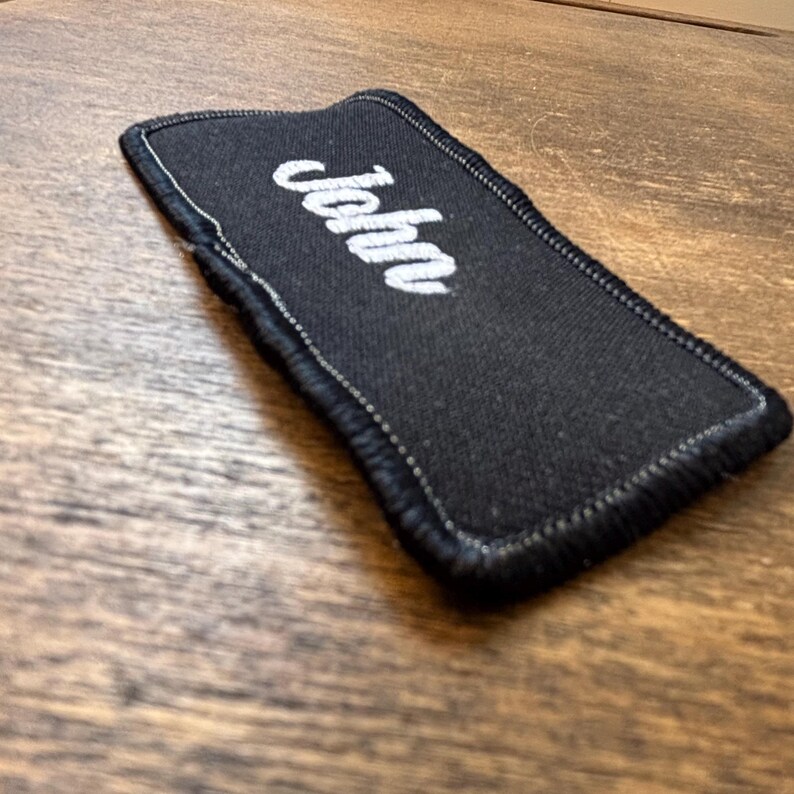 以下が含まれることがあります： 白い筆記体で「John」と刺繍された黒い長方形の携帯電話ケース。ケースには白いステッチの縁取りがあり、質感のある生地で作られています。