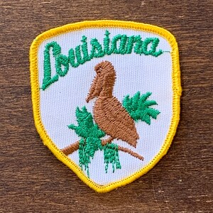 以下が含まれることがあります： 黄色い縁取りのある白い刺繍パッチ。パッチには、緑の枝に止まっている茶色のペリカンと、ペリカンの上に緑色で刺繍された「ルイジアナ」の文字が描かれています。