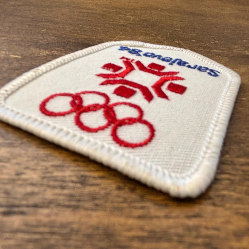 Puede incluir: Un parche blanco con detalles bordados en rojo y azul. El parche presenta los anillos ol&iacute;mpicos y el texto "Sarajevo 84". El parche tiene un borde blanco y es probablemente un recuerdo o art&iacute;culo de colecci&oacute;n.