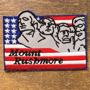 Könnte beinhalten: Gestickter Aufnäher mit Mount Rushmore und einem Design der amerikanischen Flagge. Der Aufnäher hat einen blauen Rand, mit dem Text "Mount Rushmore" in Schreibschrift.