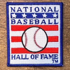 May include: A rectangular embroidered patch featuring the text NATIONAL BASEBALL HALL OF FAME TM. The design includes a blue horizontal stripe with five white stars, red and white horizontal stripes, and a central white baseball graphic.