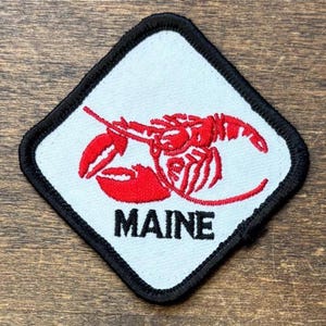 May include: A diamond-shaped embroidered patch with a white background and a thick black border. The patch features a red lobster illustration centered above the black text MAINE, which is written in a bold, capitalized sans-serif font.