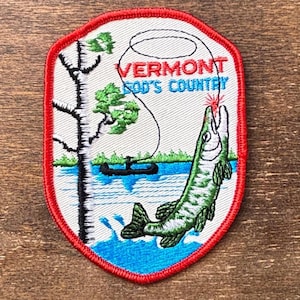 May include: Embroidered patch with a red border depicting a fishing scene. The patch shows a green fish, a tree, a boat, and the text "VERMONT GOD'S COUNTRY" in red. The background is white, with blue water and green grass.