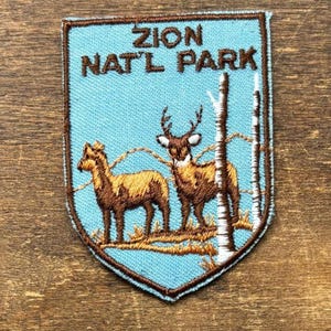 May include: Embroidered patch featuring the text "ZION NAT'L PARK" above a depiction of two deer and trees. The patch has a light blue background with brown and white details, and a brown border.