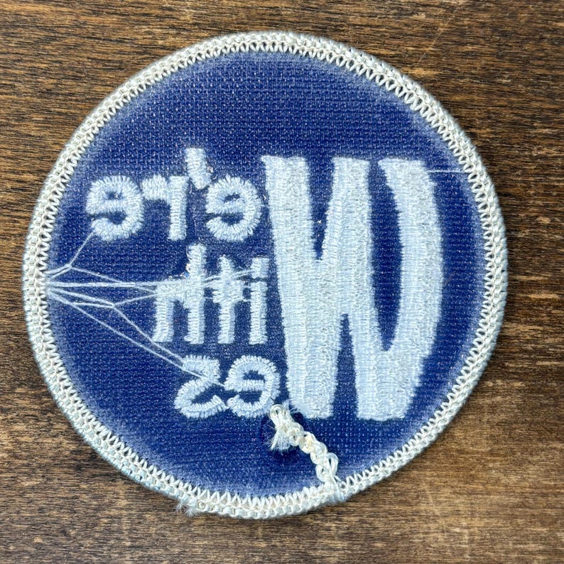 Puede incluir: Un parche bordado circular con fondo azul oscuro y borde cosido en blanco. El parche presenta el texto bordado en blanco "We're with Wes" dispuesto vertical y horizontalmente, con un gr&aacute;fico de una telara&ntilde;a en el lado izquierdo.