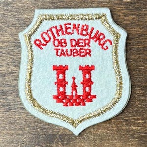 May include: A shield-shaped white fabric patch featuring red embroidered text that reads ROTHENBURG OB DER TAUBER above a red embroidered castle icon. The patch is bordered by a metallic gold thread trim along the entire outer edge.