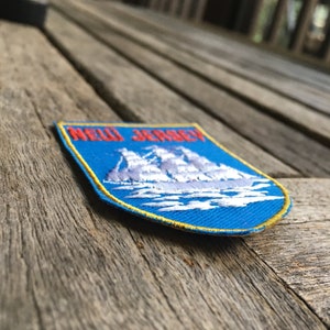 May include: A shield-shaped embroidered patch featuring a blue background with a white sailing ship graphic and the red text NEW JERSEY at the top. The patch has a yellow border and is designed for attachment to fabric items like jackets or bags.
