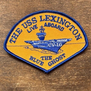 May include: A patch with a blue border and a yellow background. The patch features a blue illustration of an aircraft carrier and the text "THE USS LEXINGTON LIVE ABOARD" in blue. Below the aircraft carrier is the text "CV-16" and "THE BLUE GHOST".