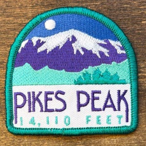 Op de afbeelding: Geborduurde patch met een berglandschap met een blauwe lucht en een witte maan. De patch leest "PIKES PEAK 14.110 FEET" in witte en turquoise letters. Het ontwerp bevat paarse bergen en groene accenten.
