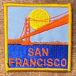 Pode incluir: Patch bordado com a Ponte Golden Gate em laranja, com um sol amarelo contra um céu azul claro. A água é azul e o texto "SAN FRANCISCO" é laranja. O patch tem uma borda amarela.