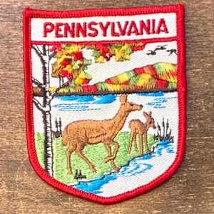 May include: A shield-shaped embroidered patch with a red border featuring the text PENNSYLVANIA in red. The design depicts a brown deer and a fawn standing near a blue stream, with a white birch tree, colorful autumn foliage, and hills.