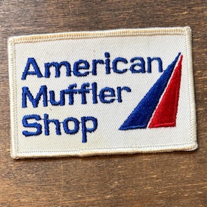 May include: Rectangular patch with the text "American Muffler Shop" in blue embroidered letters. A blue and red triangle design is on the right side. The patch is on a cream-colored background with a beige border.