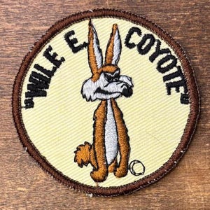 May include: A circular embroidered patch featuring a brown and white Wile E. Coyote character on a cream background. The text WILE E. COYOTE is stitched in black around the upper edge, and the patch has a dark brown stitched border.