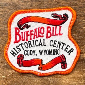 May include: Embroidered patch with an orange border and a white background. The patch features the words "Buffalo Bill Historical Center Cody, Wyoming" in red and black lettering. Two red and orange ribbons are also embroidered on the patch.
