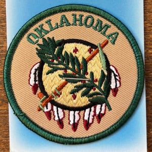 May include: Embroidered patch featuring the word "OKLAHOMA" in green lettering. The patch has a tan background with a central design of a peace pipe and olive branch, surrounded by a green border. The design includes red and white accents.
