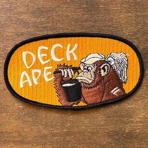 May include: An oval embroidered patch with a bright orange background and a black border. It features a brown ape wearing a white head covering, holding a black cup. The white text DECK APE is embroidered on the left side of the patch.