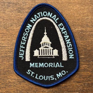 Puede incluir: Parche bordado con borde azul e interior negro. El parche presenta el texto "Jefferson National Expansion Memorial St. Louis, MO." en blanco y una ilustración blanca de un edificio.