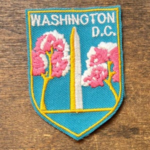 May include: Embroidered patch featuring the text "WASHINGTON D.C." in white. The patch has a shield shape with a light blue background and a yellow border. The design includes a white monument and pink and white flowering trees.