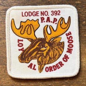 Può includere: Una toppa ricamata quadrata con sfondo bianco e bordo cucito. Presenta un'illustrazione di una testa di alce marrone e gialla con testo rosso che recita LODGE NO. 392, P.A.P. e LOYAL ORDER OF MOOSE disposto attorno al grafico centrale.