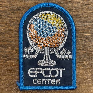 May include: A navy blue embroidered patch featuring the Epcot Center logo. The logo depicts a stylized geodesic sphere with yellow, orange, and blue segments. The patch has a blue border and the words "EPCOT CENTER" in silver lettering.