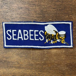 May include: A rectangular embroidered patch with a blue background and a white border. The text SEABEES is stitched in white capital letters on the left. On the right, a cartoon bee wearing a white sailor hat holds a yellow tool.