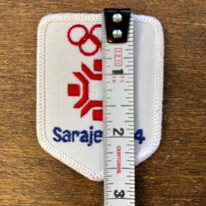 Puede incluir: Un parche blanco con detalles bordados en rojo y azul. El parche presenta los anillos ol&iacute;mpicos, una figura estilizada y la palabra "Sarajevo 84". El parche mide aproximadamente 5 cm de altura.