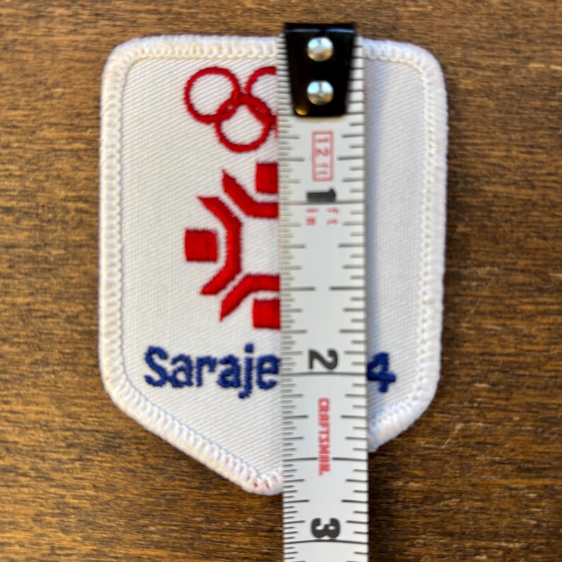 Puede incluir: Un parche blanco con detalles bordados en rojo y azul. El parche presenta los anillos ol&iacute;mpicos, una figura estilizada y la palabra "Sarajevo 84". El parche mide aproximadamente 5 cm de altura.