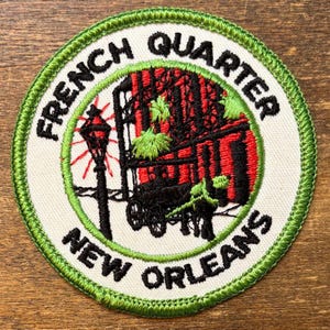 May include: A circular embroidered patch with a bright green stitched border on a white background. It features a black silhouette of a horse-drawn carriage and a street lamp, with red and green accents. The text reads French Quarter New Orleans.