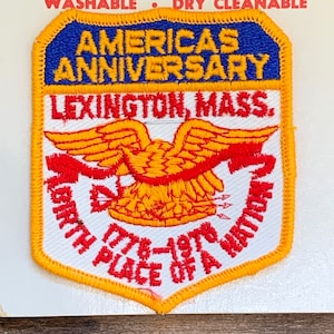 Puede incluir: Un parche bordado blanco, rojo y amarillo con el texto "America's Anniversary, Lexington, Mass. Birth Place of a Nation 1776-1976". El parche presenta un águila roja y amarilla con flechas.