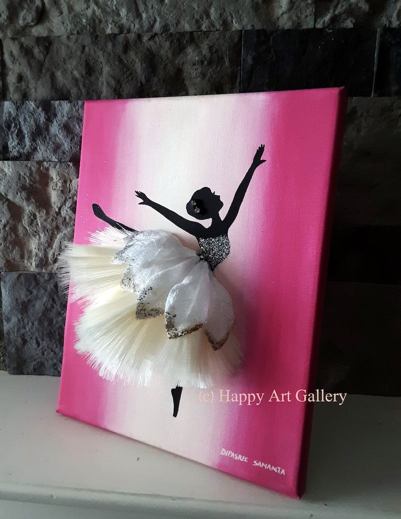 May include: A pink and white ombre canvas painting of a ballerina silhouette with a white tulle tutu. The tutu is embellished with gold glitter and white feathers. The painting is signed "Happy Art Gallery" and "Dyvasree Samanta".
