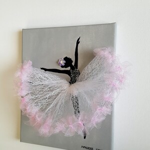 May include: A gray canvas painting of a ballerina silhouette with a pink tulle tutu. The tutu is layered and has a pink trim. The ballerina has a purple flower on her head. The painting is signed "Dipasree Samantha (c) Happy Art Gallery".