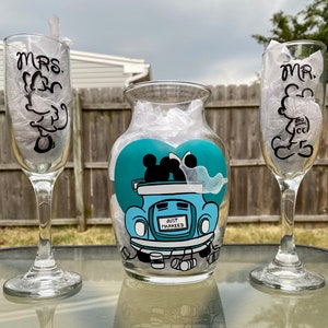 Op de afbeelding: Een bruiloft centerpiece met een heldere glazen vaas met een turquoise hartontwerp. Het hart heeft een cartoon afbeelding van Mickey en Minnie Mouse in een auto met de woorden "Just Married". Twee heldere glazen champagnefluiten met zwarte letters die "Mrs." en "Mr." zeggen, staan aan weerszijden van de vaas.