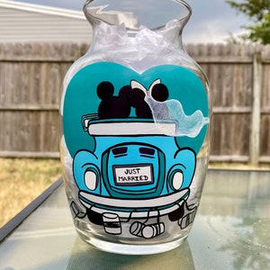 May include: A clear glass vase with a teal heart design featuring Mickey and Minnie Mouse in a blue car with the words "Just Married".
