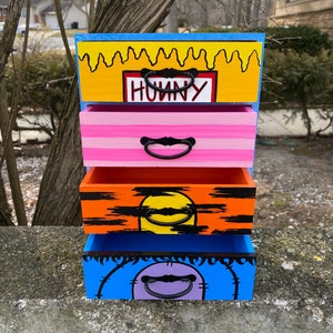 Può includere: Una cassettiera in legno a quattro cassetti dipinta con colori vivaci. Il cassetto superiore è giallo con vernice nera che cola e la parola "HONEY" in un rettangolo rosso. Il secondo cassetto è rosa con strisce bianche. Il terzo cassetto è arancione con strisce nere e un cerchio giallo. Il cassetto inferiore è blu con un cerchio viola.