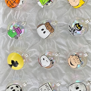 May include: A selection of clear glass baubles, each decorated with cartoon characters. The designs include Charlie Brown, Snoopy, and the Great Pumpkin, with the text "Welcome Great Pumpkin" on one of the baubles.