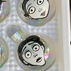 May include: Set of four glass ornaments featuring characters from the movie The Nightmare Before Christmas. The ornaments are white with black and white painted details. The characters include Jack Skellington, Sally, and two other characters.