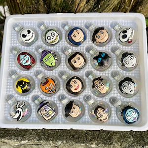 May include: A set of 18 clear glass ornaments with various characters from the Nightmare Before Christmas and Corpse Bride films. The ornaments are arranged in a white plastic tray.