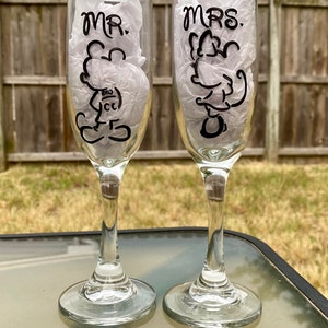 May include: Two clear glass champagne flutes with black painted designs. The left flute says "MR." with a cartoon Mickey Mouse design. The right flute says "MRS." with a cartoon Minnie Mouse design.