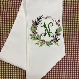 May include: White kitchen towel with an embroidered wreath of pine branches, pine cones, and berries with the letter "N" in the center.
